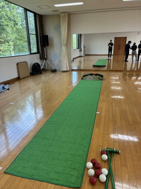 Indoor gym or studio with a long green putting mat extending toward a mirror; balls and cues/tools are at the near end, and people stand in the background.