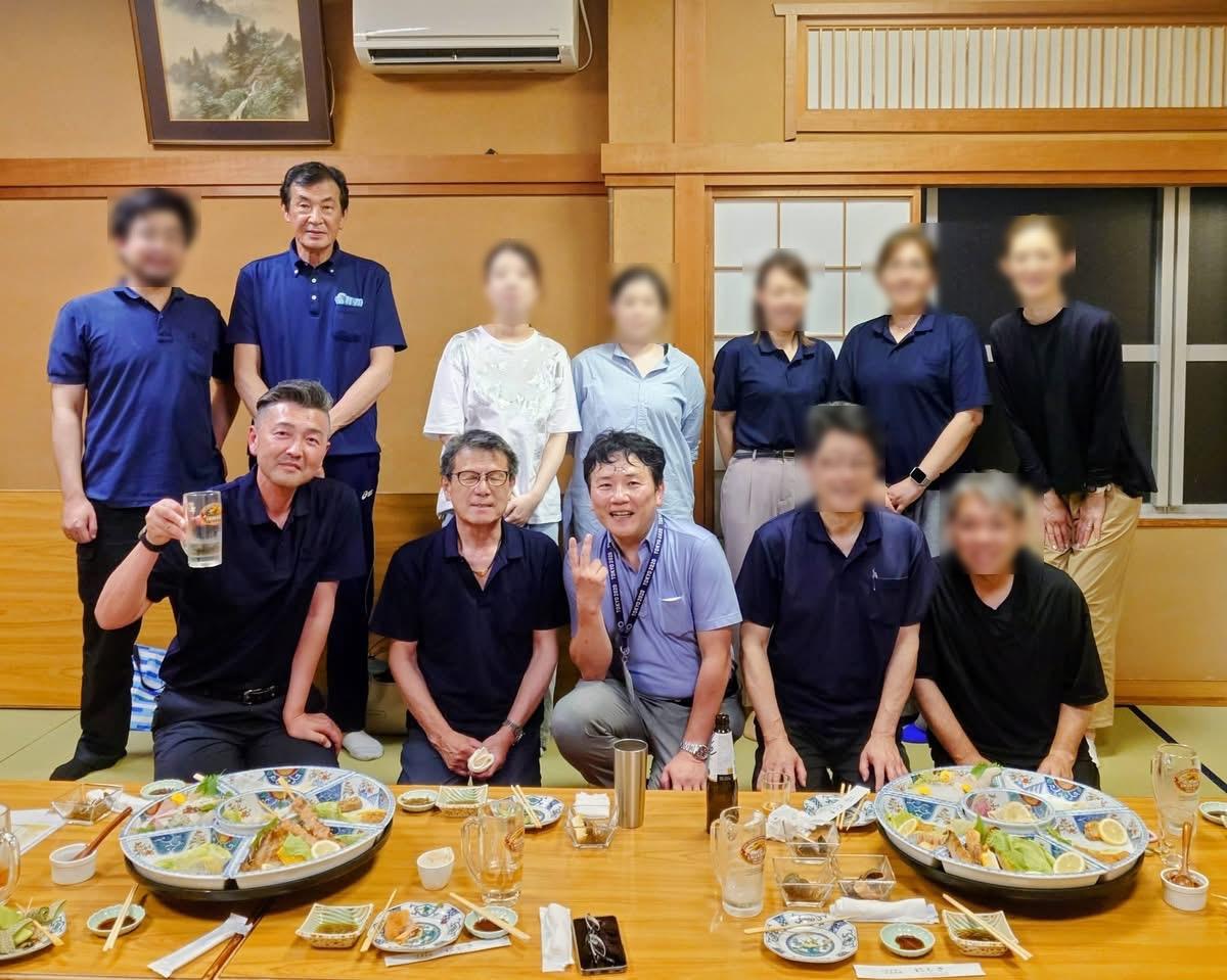 Group of coworkers posing for a photo around a low dining table with plates of food and drinks in a warm Japanese-style room (faces lightly blurred). - Includes the sense of a celebratory meal in a traditional setting.